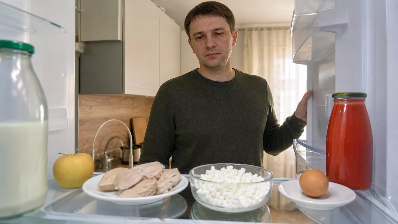 Мужчина рассматривает в холодильнике творог, мясо, яйцо, томатный сок, яблоко и кефир